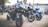 Several high-performance motorcycles are parked together in an outdoor area. The motorcycles are predominantly black with accents of green, blue, and silver. In the background, a few people are standing, and more motorcycles are visible.