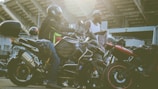 Several motorcyclists gather near their parked motorcycles in an urban setting with a large structure in the background. One person is seated on a motorcycle wearing a helmet and protective gear, while another stands nearby in a casual outfit holding a piece of clothing.