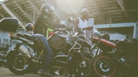 Several motorcyclists gather near their parked motorcycles in an urban setting with a large structure in the background. One person is seated on a motorcycle wearing a helmet and protective gear, while another stands nearby in a casual outfit holding a piece of clothing.