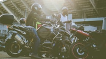 Several motorcyclists gather near their parked motorcycles in an urban setting with a large structure in the background. One person is seated on a motorcycle wearing a helmet and protective gear, while another stands nearby in a casual outfit holding a piece of clothing.