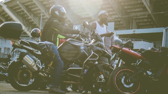 Several motorcyclists gather near their parked motorcycles in an urban setting with a large structure in the background. One person is seated on a motorcycle wearing a helmet and protective gear, while another stands nearby in a casual outfit holding a piece of clothing.