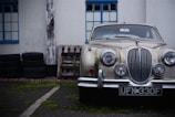 A classic vintage car with a distinctively designed front grille and round headlights is parked next to a stack of tires and wooden pallets. The car shows signs of aging with moss or dirt buildup on its surface. The setting appears to be urban, with the vehicle positioned in front of a building featuring blue-framed windows. The atmosphere is somewhat nostalgic and rustic.