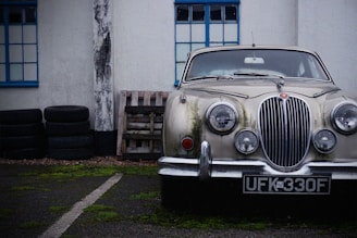 A classic vintage car with a distinctively designed front grille and round headlights is parked next to a stack of tires and wooden pallets. The car shows signs of aging with moss or dirt buildup on its surface. The setting appears to be urban, with the vehicle positioned in front of a building featuring blue-framed windows. The atmosphere is somewhat nostalgic and rustic.