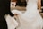 Two people appear to be part of a wedding ceremony. One is wearing a white wedding dress with lace details, while the other, dressed in black attire, is assisting by holding the dress. The background is blurred, suggesting an outdoor setting.