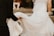 Two people appear to be part of a wedding ceremony. One is wearing a white wedding dress with lace details, while the other, dressed in black attire, is assisting by holding the dress. The background is blurred, suggesting an outdoor setting.