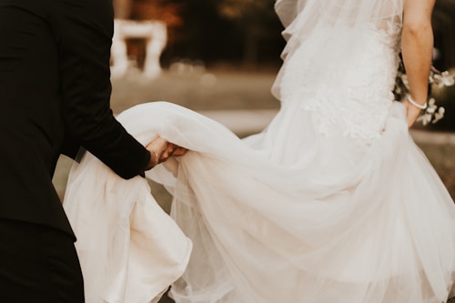 Two people appear to be part of a wedding ceremony. One is wearing a white wedding dress with lace details, while the other, dressed in black attire, is assisting by holding the dress. The background is blurred, suggesting an outdoor setting.