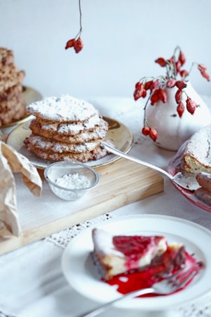 A tabletop setting with a plate of stacked cookies dusted with powdered sugar, a small bowl containing more powdered sugar, and a slice of cake with a vibrant red sauce on a plate. A rustic white vase holds red berries as decoration, adding to the cozy and inviting atmosphere. The scene is set on a wooden board and lace tablecloth, creating a homely and warm feel.