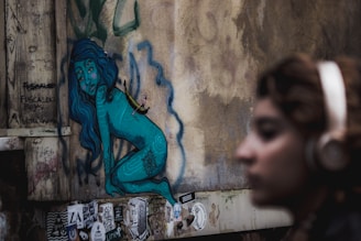 A street art mural on a wall depicts a blue humanoid figure, with long hair, and intricate patterns on its body. The figure appears to sit or squat against a textured, weathered wall. Various stickers and graffiti surround the artwork, adding to an urban aesthetic. In the foreground, a person with blurred features and wearing headphones stands out of focus, contributing to the scene's depth.