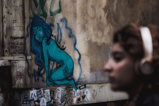 A street art mural on a wall depicts a blue humanoid figure, with long hair, and intricate patterns on its body. The figure appears to sit or squat against a textured, weathered wall. Various stickers and graffiti surround the artwork, adding to an urban aesthetic. In the foreground, a person with blurred features and wearing headphones stands out of focus, contributing to the scene's depth.