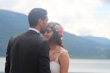 A man and a woman are embracing outdoors with a backdrop of mountains and a lake. The man is dressed in a black suit and white shirt while the woman is wearing a white dress with lace details and a colorful flower crown. They appear to be in a happy and intimate moment, with the man gently kissing the woman on her forehead.