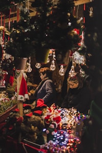 A festive market stall filled with Christmas decorations and holiday lights. Two people are browsing through various ornaments and small Santa figures. Garland and pine trees are decorated with small lights, contributing to the cozy, festive atmosphere.