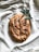 A loaf of rustic bread with a golden brown crust, lightly dusted with coarse salt. The bread rests on a soft white fabric, alongside a sprig of olive leaves, giving a natural and artisanal presentation.