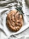 A loaf of rustic bread with a golden brown crust, lightly dusted with coarse salt. The bread rests on a soft white fabric, alongside a sprig of olive leaves, giving a natural and artisanal presentation.