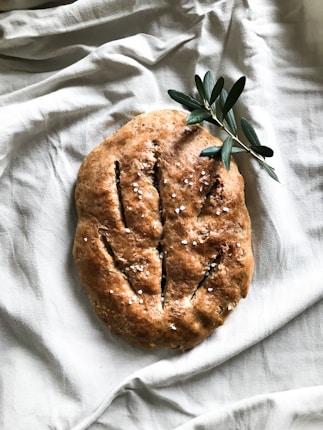A loaf of rustic bread with a golden brown crust, lightly dusted with coarse salt. The bread rests on a soft white fabric, alongside a sprig of olive leaves, giving a natural and artisanal presentation.