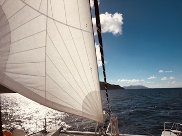 A large white sail is prominently displayed on a sailboat cruising through the ocean. The sun reflects off the water, creating a shimmering effect, while distant mountains and a clear blue sky form the backdrop.