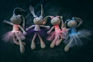 Four crocheted bunny dolls wearing tulle skirts in different colors are arranged side by side on a dark, textured surface. Each bunny has large ears and a bow on its head.