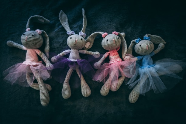 Four crocheted bunny dolls wearing tulle skirts in different colors are arranged side by side on a dark, textured surface. Each bunny has large ears and a bow on its head.