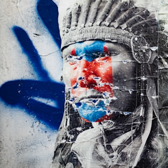 A weathered street art poster features a monochrome image of a person wearing a traditional headdress. The person's face is partially covered in vibrant blue and red paint. The poster is peeling, revealing the rough texture of the surface it is pasted on. Blue graffiti handprints appear in the background, adding to the layered, urban aesthetic.