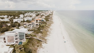A coastal aerial view features a long, sandy beach stretching alongside a residential area with modern and traditional houses. The water appears calm, with a few people relaxing under umbrellas on the beach. Lush greenery separates the houses from the shoreline, and the sky is partly cloudy.