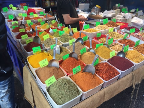 A market stall displays a variety of colorful spices in rectangular containers. Each container is filled with a different spice, and metal scoops are placed in them. Brightly colored labels are attached to each spice, indicating their names. A person is partially visible behind the stall, possibly attending to customers. The spices are arranged in a neat, organized fashion, creating an appealing visual display.