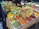 A market stall displays a variety of colorful spices in rectangular containers. Each container is filled with a different spice, and metal scoops are placed in them. Brightly colored labels are attached to each spice, indicating their names. A person is partially visible behind the stall, possibly attending to customers. The spices are arranged in a neat, organized fashion, creating an appealing visual display.