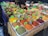 A market stall displays a variety of colorful spices in rectangular containers. Each container is filled with a different spice, and metal scoops are placed in them. Brightly colored labels are attached to each spice, indicating their names. A person is partially visible behind the stall, possibly attending to customers. The spices are arranged in a neat, organized fashion, creating an appealing visual display.