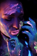 A person with eyes closed, their face and hands covered in vibrant neon paint. The colors are illuminated under blacklight, creating a surreal and artistic effect.