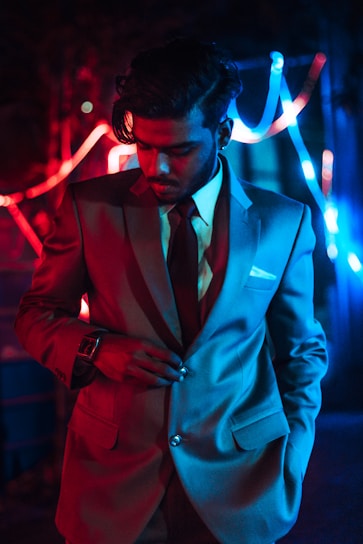 A person wearing a suit stands under colorful, dramatic lighting, which casts red and blue hues on their clothes and face. The individual appears to be adjusting their suit while looking downward. The background is dimly lit and features abstract neon lights, adding a modern and stylish feel to the scene.
