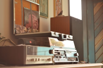 A vintage audio setup featuring a turntable and speakers is situated on a wooden surface. Several framed artworks and photographs are displayed on the wall above, adding a personal and nostalgic touch to the scene. The image conveys a sense of a cozy, retro-themed interior.