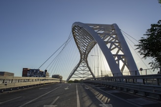 An arched steel bridge with a modern design stretches over a road. The architecture features cables connected to the main arch, creating geometric patterns. Surrounding the bridge are a few buildings, and trees are visible to the side. The lighting suggests it is early morning or late afternoon, with the sun creating shadows and a calm atmosphere.