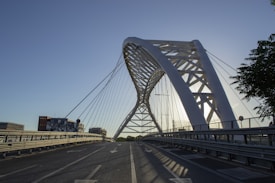 An arched steel bridge with a modern design stretches over a road. The architecture features cables connected to the main arch, creating geometric patterns. Surrounding the bridge are a few buildings, and trees are visible to the side. The lighting suggests it is early morning or late afternoon, with the sun creating shadows and a calm atmosphere.