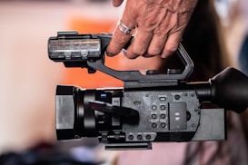 A close-up of a professional video camera being operated by a hand, emphasizing the camera's controls and dials. The background is softly blurred, with subtle hints of color, creating a focused view on the camera equipment.