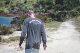 A person wearing a dark long-sleeved shirt with 'Stussy Designs, Laguna Beach, CA' printed on the back walks along a dirt path. The background features greenery and a body of water.