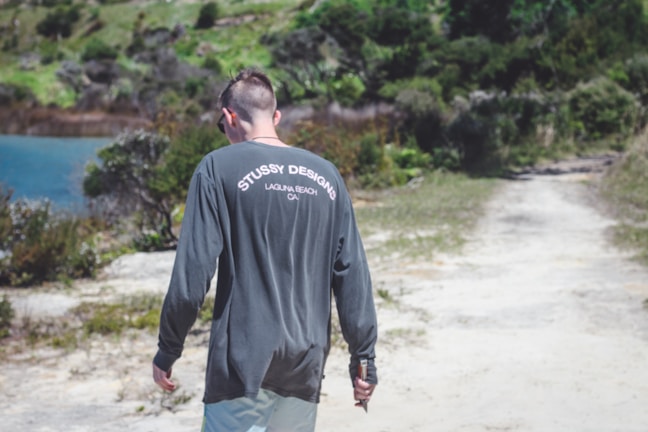 A person wearing a dark long-sleeved shirt with 'Stussy Designs, Laguna Beach, CA' printed on the back walks along a dirt path. The background features greenery and a body of water.