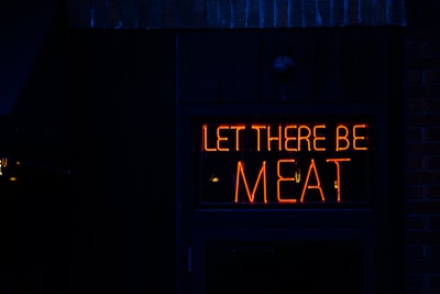 A neon sign with the words 'LET THERE BE MEAT' displayed in bright red lights against a dark background.