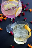 Two glasses of cocktails are placed on a dark surface adorned with scattered colorful flower petals. The first glass contains a pink cocktail garnished with lemon slices and a small sprig of flowers. The second glass holds a clear cocktail with ice and is garnished with a lemon peel.