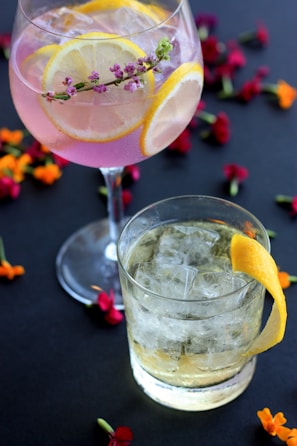 Two glasses of cocktails are placed on a dark surface adorned with scattered colorful flower petals. The first glass contains a pink cocktail garnished with lemon slices and a small sprig of flowers. The second glass holds a clear cocktail with ice and is garnished with a lemon peel.