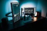 A modern computer setup featuring two monitors; one vertical and the other horizontal. The workspace includes a keyboard, mouse, speakers, headphones, and a small desk lamp. The room is dimly lit with a cool blue tone illuminating the area from the lamp and the screens.