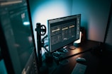 A modern and dimly lit workspace featuring a computer setup with dual monitors. The main monitor displays a graphic design project with text, while a keyboard and mouse are placed on the desk. Mood lighting is provided by a small lamp, casting a soft glow over the workspace.