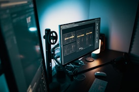 A modern and dimly lit workspace featuring a computer setup with dual monitors. The main monitor displays a graphic design project with text, while a keyboard and mouse are placed on the desk. Mood lighting is provided by a small lamp, casting a soft glow over the workspace.