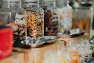 A variety of glass jars filled with different types of cookies and snacks are lined up on a wooden counter. The jars are clear and labeled, showing an assortment of cookies, including chocolate chip and what appears to be other baked goods. Metal scoops are placed nearby, suggesting a self-service setup.