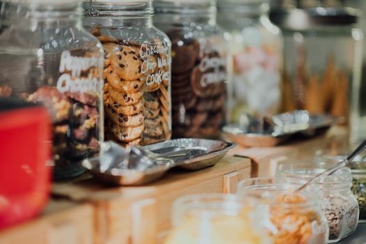 A variety of glass jars filled with different types of cookies and snacks are lined up on a wooden counter. The jars are clear and labeled, showing an assortment of cookies, including chocolate chip and what appears to be other baked goods. Metal scoops are placed nearby, suggesting a self-service setup.
