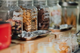 A variety of glass jars filled with different types of cookies and snacks are lined up on a wooden counter. The jars are clear and labeled, showing an assortment of cookies, including chocolate chip and what appears to be other baked goods. Metal scoops are placed nearby, suggesting a self-service setup.