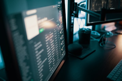 Several computer monitors display various screens with digital content, including text and graphics. A keyboard is visible on the desk, along with a smart speaker and some cables. The lighting is dim, suggesting a work or gaming setup.