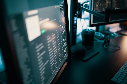 Several computer monitors display various screens with digital content, including text and graphics. A keyboard is visible on the desk, along with a smart speaker and some cables. The lighting is dim, suggesting a work or gaming setup.