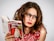 A woman with long brown hair and glasses is wearing a red dress with a white floral pattern. She is holding an open book titled 'Ça peut pas rater!' by Gilles Legardinier, which has a cover featuring a white cat.