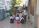 A group of young children is seated on small stools outdoors in an alleyway, attentively engaged with an adult woman standing nearby who appears to be teaching or talking to them. The scene is surrounded by various elements such as potted plants, a water cooler, parked motorcycles, and an arrangement of colorful items in the background. The children are wearing matching uniforms with white shirts and dark overalls.