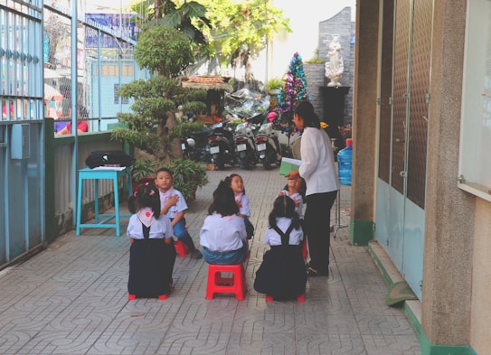 A group of young children is seated on small stools outdoors in an alleyway, attentively engaged with an adult woman standing nearby who appears to be teaching or talking to them. The scene is surrounded by various elements such as potted plants, a water cooler, parked motorcycles, and an arrangement of colorful items in the background. The children are wearing matching uniforms with white shirts and dark overalls.