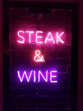 Neon sign with the words 'STEAK & WINE' glowing in pink and purple against a dark brick wall background.