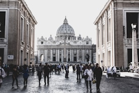 A busy street scene with people walking on a cobblestone path flanked by two large architectural buildings. A grand domed structure, possibly St. Peter's Basilica, is visible in the background, with an obelisk in front of it. The area is lively, with many individuals of varying ages and appearances engaging in activities. The lighting suggests a daytime setting.